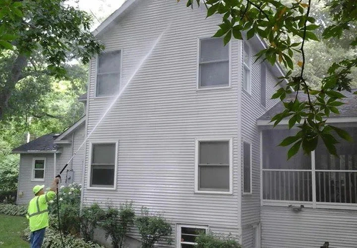 Professional technician performing soft wash vinyl siding cleaning on a grey two-story house.